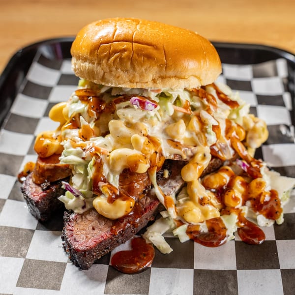Brisket sandwich with coleslaw, macaroni and cheese, and BBQ sauce on a checkered paper-lined tray.