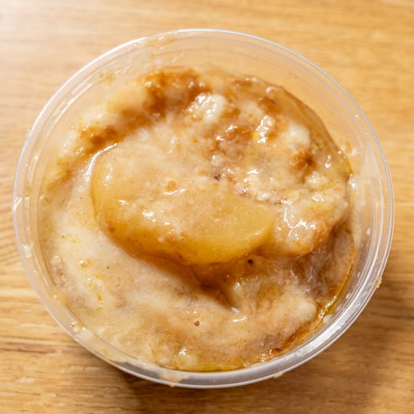 Close-up of a clear plastic container filled with a creamy, light brown dessert with fruit chunks.