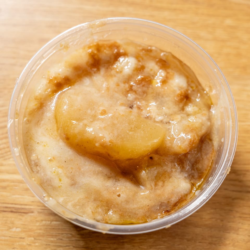 Close-up of a clear plastic container filled with a creamy, light brown dessert with fruit chunks.