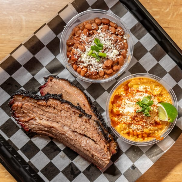 Smoked brisket slices, a cup of pinto beans, and a cup of elote on a checkered tray.