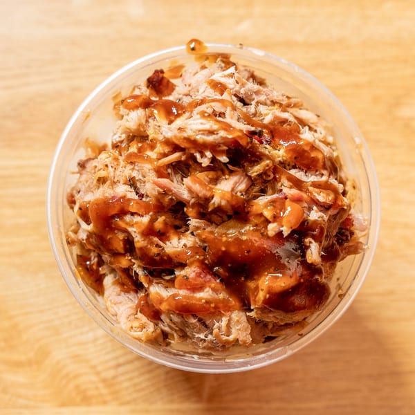 Close-up of a clear plastic cup filled with pulled pork drizzled with barbecue sauce.