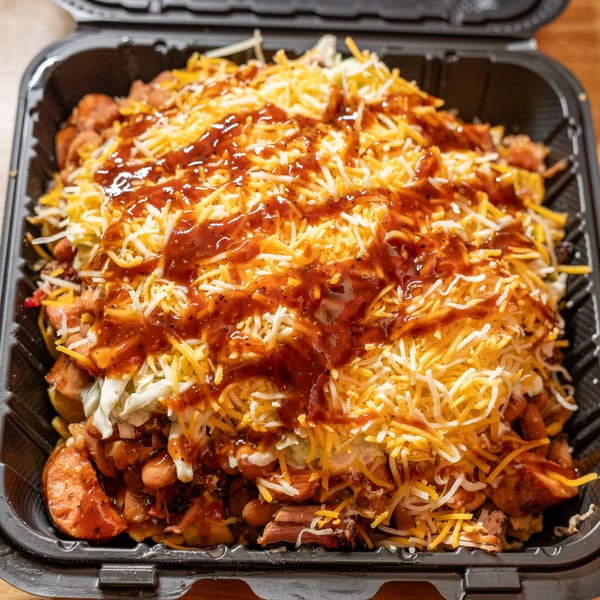 Loaded BBQ dish in a black takeout container, topped with shredded cheese and barbecue sauce.